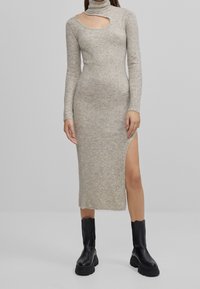 Robe en maille beige avec un col montant, présentant une découpe sur l'épaule gauche et une fente latérale, associée à des bottes noires à semelles épaisses.