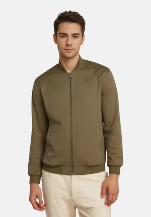 Jeune homme portant une veste zippée vert olive avec des poignets côtelés et un pantalon beige, debout devant un fond clair uni.