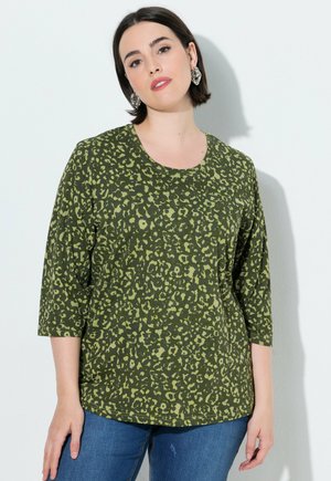 Femme aux cheveux courts et foncés portant un haut vert à imprimé léopard avec des manches trois quarts et un jean bleu, debout devant un fond clair.