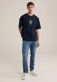 Mornarsko modra bombažna majica s kratkimi rokavi, ki ima kvadratno grafiko z napisom "BLUE RIDGE". Ujemajoča svetlo modra kavbojka in bele superge.