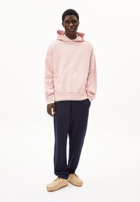 Felpa in cotone rosa chiaro con tasca frontale e cappuccio con coulisse, abbinata a pantaloni blu navy e scarpe beige slip-on.