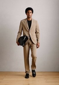 Hombre caminando por dentro llevando un traje beige, camisa negra, zapatos negros y llevando una bolsa texturizada negra en su mano izquierda.