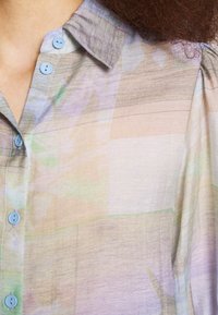 Blusa transparente con una paleta de colores pastel y patrones abstractos, un cuello puntiagudo y botones azul claro en el frente.