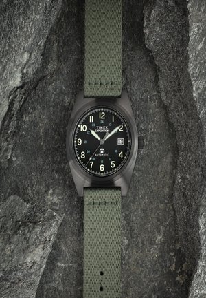 Zegarek Timex Expedition z matową czarną kopertą, czarną tarczą, białymi cyframi i zielonym materiałowym paskiem. Posiada mechanizm automatyczny oraz wyświetlacz daty.