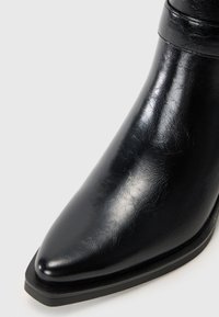 Botín de cuero negro hasta el tobillo con punta afilada, textura elegante y suela negra baja. Presenta una sutil banda en el eje para detalle de diseño.