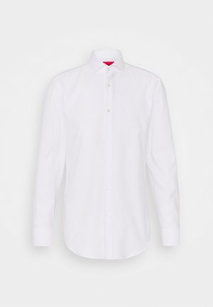 Camisa blanca de algodón con mangas largas, cuello abotonado y dobladillo curvo. Presenta botones claros y un acento rojo en el interior del cuello.