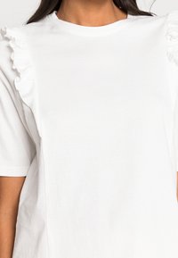 Bílá krátká tričko s rukávy zdobenými volánky. Vyrobeno z měkké tkaniny, s uvolněným střihem a kulatým výstřihem.