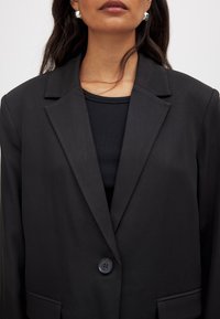 Mustaline musta pintsak ühe nööbiga, sälkpingutustega ja siledast materjalist. Kanda musta ribakujulise topi üle, mis on sobitatud disainiga.