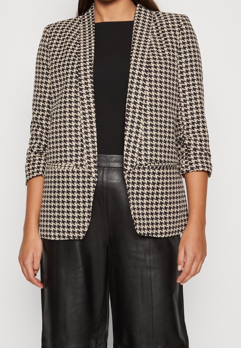 Femme portant un top noir, un pantalon en cuir noir et un blazer beige et noir à motif pied-de-poule avec manches retroussées.