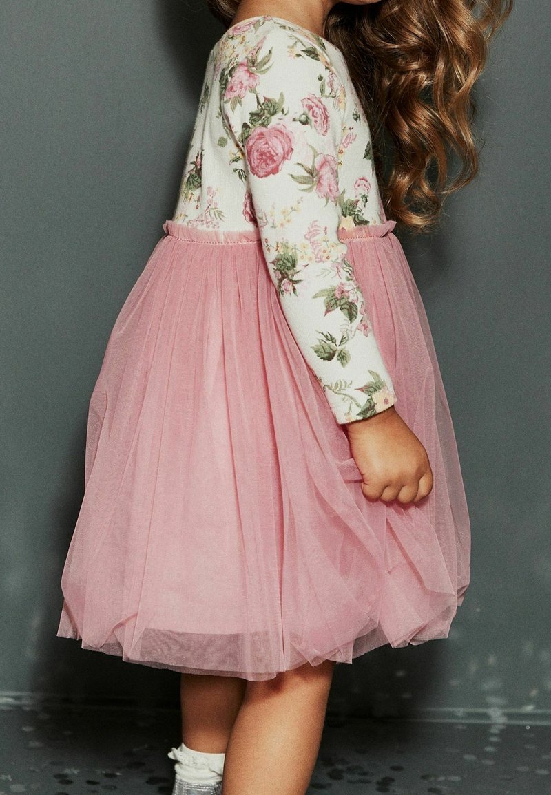 Enfant portant un haut à manches longues fleuri et une jupe en tulle rose, se tenant de côté avec des cheveux bouclés devant un fond gris uni.