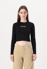 Crna dugosleeve skraćena majica s "HUGO" logotipom u bijeloj boji na prsima, u kombinaciji s svijetlosmešanim hlačama. Glatka tkanina, prilagođen dizajn.
