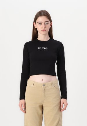Black long-sleeve cropped top with "HUGO" logo in white on the chest, paired with light beige pants. Smooth fabric, fitted design.