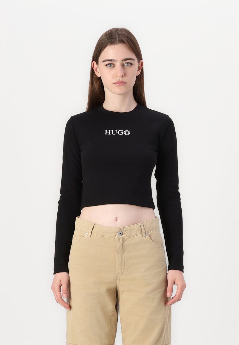Crna dugosleeve skraćena majica s "HUGO" logotipom u bijeloj boji na prsima, u kombinaciji s svijetlosmešanim hlačama. Glatka tkanina, prilagođen dizajn.