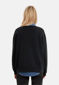 Blonde person wearing a black sweatshirt over a blue denim shirt and black pants, standing facing away against a plain background.