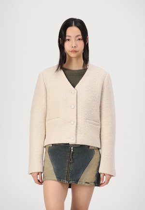 Giacca beige, strutturata, corta con scollo a V, bottoni frontali e tasche laterali, abbinata a una gonna in denim multicolore con zip.