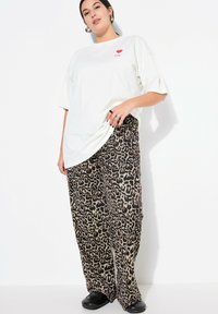 T-shirt bianca oversize con grafica a cuore rossa. Pantaloni larghi con stampa leopardata in beige e marrone. Scarpe nere slip-on. Modello in piedi contro uno sfondo bianco.