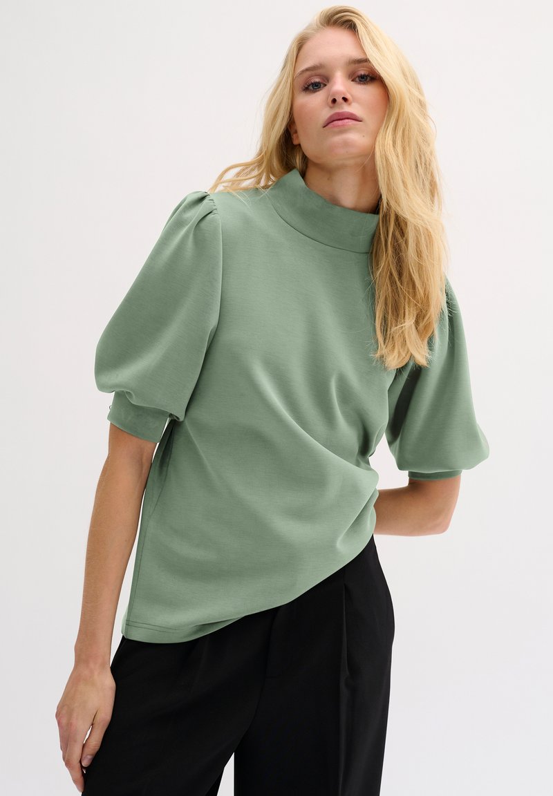 Maglietta verde chiaro a maniche corte con colletto alto e maniche a sbuffo, realizzata in un materiale liscio. Abbinata a pantaloni neri.