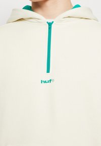 Sweat à capuche crème avec une fermeture éclair turquoise, doté d'une poche kangourou et du logo brodé "huf" assorti en turquoise sur la poitrine.