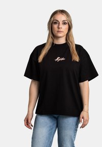 Černé oversized tričko s růžovým logem na hrudi. Bavlněná látka má hladkou texturu a krátké rukávy.