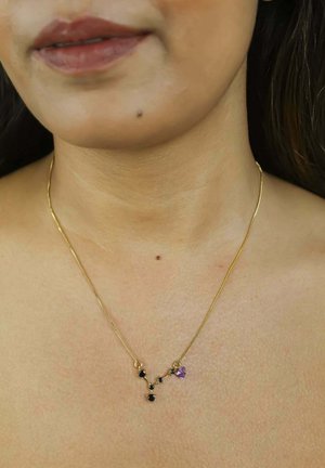 Gros plan sur le cou d'une personne portant un collier constellation en or avec des pierres noires et un petit pendentif cœur violet.