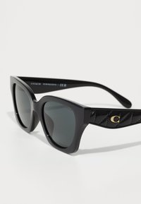 Lunettes de soleil noires en forme de chat avec des verres gris et une finition brillante. Présente des branches noires texturées avec un accent logo doré.