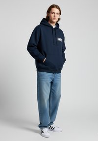 Felpa blu navy con "AMOUR" in testo bianco, tasca a marsupio, abbinata a jeans azzurri e sneakers bianche con strisce nere.