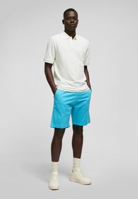 Witte polo met korte mouwen, gecombineerd met felblauwe shorts. Model draagt crèmekleurige sneakers en enkelsokken. Eenvoudig, casual ontwerp.