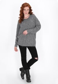 Grauer, gestrickter Pullover mit lockerer Passform, überschnittenen Schultern und fransigem Saum, kombiniert mit schwarzen, zerrissenen Jeans und schwarzen Stiefeletten.