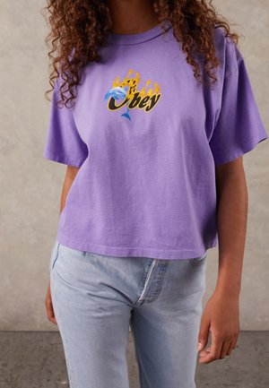 Frau trägt ein lila, bauchfreies T-Shirt mit einem grafischen Bild eines brennenden Delfins und dem Wort "Obey", kombiniert mit hellblauen Jeans.