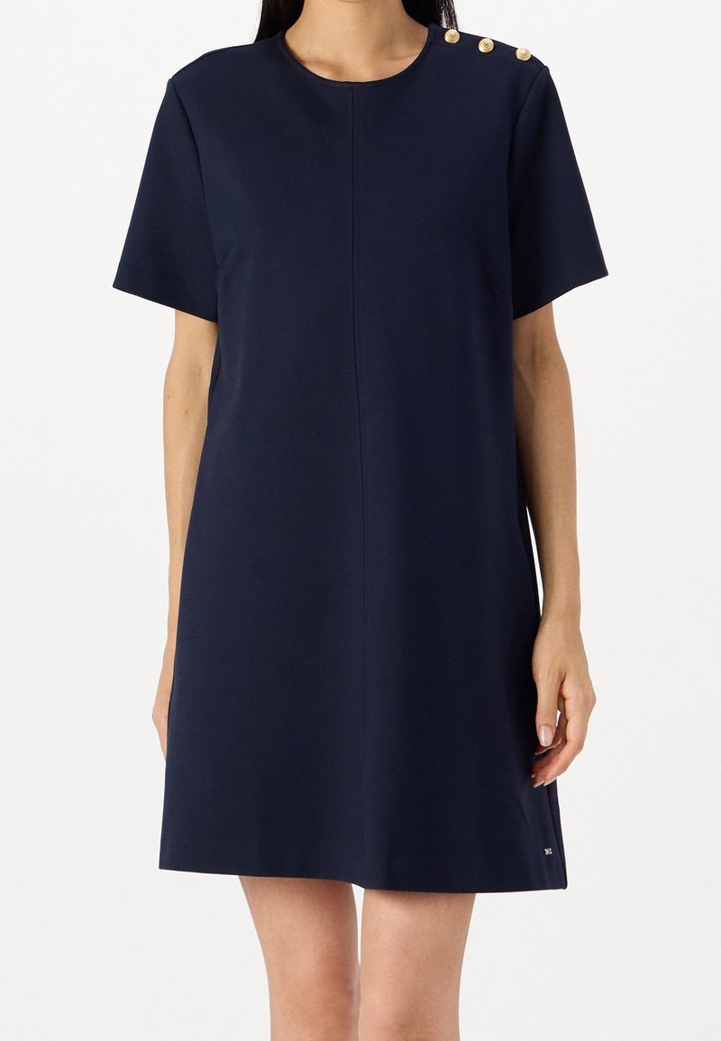 Femme portant une robe bleu marine à manches courtes avec trois boutons dorés sur l'épaule gauche et un col rond.