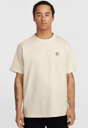 Man with short curly hair wearing a beige t-shirt with a small circular logo on the chest and tattoos visible on both forearms.