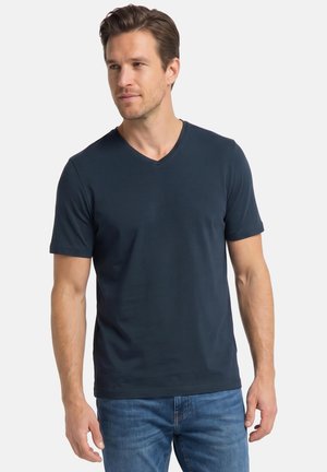 Mann trägt ein dunkelblaues V-Ausschnitt-T-Shirt mit kurzen Ärmeln und blaue Jeans, steht mit neutralem Gesichtsausdruck, Hände entspannt an den Seiten.