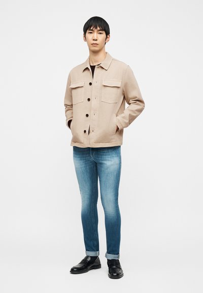 Άνδρας στέκεται με τα χέρια στις τσέπες ενός μπεζ μπουφάν, φορά ξεθωριασμένο μπλε τζιν με γυρισμένα μανίκια στους αστραγάλους και μαύρα δερμάτινα παπούτσια χωρίς κορδόνια σε λευκό φόντο.