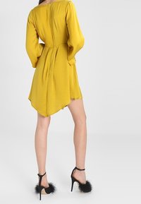 Robe jaune, longueur genou, avec manches longues et taille cintrée. Présente un ourlet arrondi et un tissu texturé. Sandales noires à talons avec des accents en fourrure.
