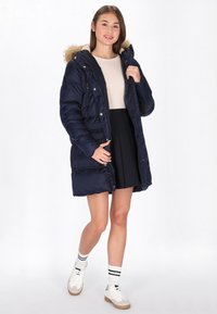 Marineblå pufferjakke med en hætte i pels, lynlås lukning, forlommer og en quiltet design, båret over en lys top og sorte nederdel.