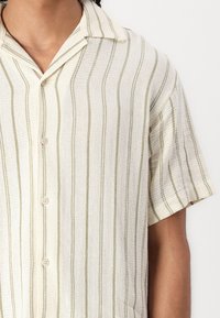 Camicia a maniche corte con bottoni realizzata in tessuto testurizzato. Base crema chiaro con strisce verticali color verde oliva e una tasca sul petto.