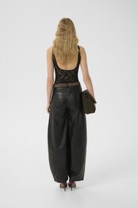 Woman with long wavy blonde hair wearing black lace sleeveless top, wide-leg black leather pants, holding a dark clutch, standing on white background.