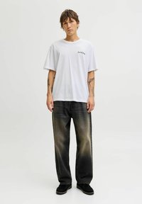 Camiseta de algodón blanca de manga corta con un estampado en el pecho izquierdo. Combinada con jeans de mezclilla oscuros y de corte holgado con un efecto degradado.