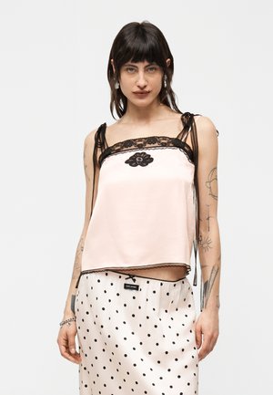 Giovane con capelli scuri indossa una canottiera rosa pallido con rifiniture in pizzo nero e una gonna a pois, su uno sfondo chiaro e uniforme.