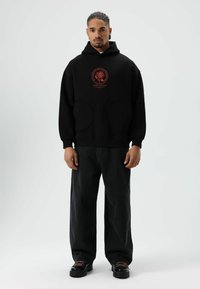 Zwarte hoodie met rode grafische print, grote zakken aan de voorkant, geribbelde manchetten en zwarte wijde jeans, gecombineerd met zwarte schoenen met een dikke zool.