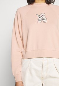 Ljust rosa sweatshirt med raglanärmar, prydd med en tryckt design av två tecknade kaniner som håller en "Levi's" -banner. Mjuk textur.