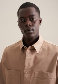 Camicia button-up marrone chiaro con colletto strutturato, due tasche frontali e tessuto di cotone liscio. Presenta dettagli di cucitura sottili.