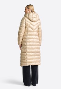 Beige gewatteerde lange jas met een capuchon, voorzien van een chevronpatroon, glanzende textuur en ritszakken. Gepaarde met losse zwarte broeken.