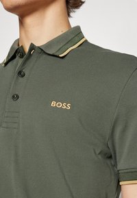 Mörkgrön polo med en texturerad tyg. Den har en krage med ett grönt och guldigt randmönster och en guld "BOSS" logga på bröstet.