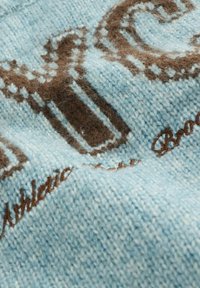 Hellblaue Strickware mit brauner Schrift und Mustern. Weiche Textur mit leicht fuzzy Erscheinung und aufwendigen Stickdetails.