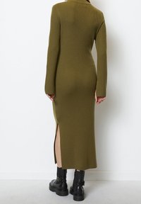 Robe en tricot vert olive avec col montant, manches longues, fentes latérales et texture côtelée, associée à des bottes noires épaisses.