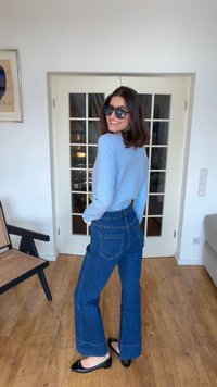 Femme portant des lunettes de soleil, un pull bleu clair, un jean bleu foncé et des ballerines noires, debout à l'intérieur sur un sol en bois près de portes vitrées.