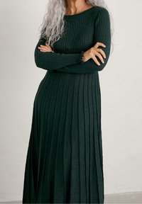 Abito lavorato a maglia verde scuro con texture a coste, maniche lunghe e polsini con volant. L'abito presenta una silhouette svasata e un design lungo fino al pavimento.