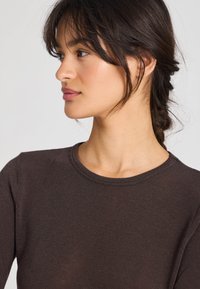 Cămașă maro cu mâneci lungi, dantelată, cu guler rotund. Textură moale, croială ajustată, cu cusături vizibile și detalii minimale.