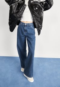 Černá lesklá péřová bunda s bílými pruhy, vysoké modré džíny s rozšířenými nohavicemi a béžové tenisky na modrém pozadí.